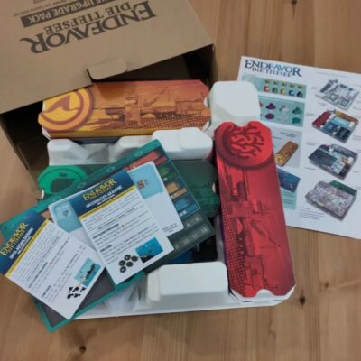 Karton mit Brettspiel "Endeavor: Die Tiefsee" auf Holztisch; offene Schachtel zeigt farbige Spielkomponenten und Anleitungen. Daneben liegt ein Übersichtspapier mit Spielgrafiken und Erklärungen.