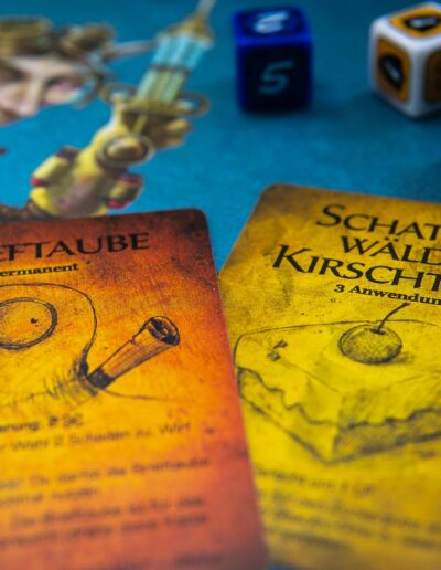 Nahaufnahme von zwei Spielkarten auf einem Tisch. Die linke Karte zeigt eine Zeichnung einer Brieftaube, darunter steht „Brieftaube Permanent“. Die rechte Karte zeigt ein Stück Kirschkuchen mit der Aufschrift „Schattenwälder Kirschtorte 3 Anwendungen“. Im Hintergrund sind Würfel zu sehen, die Zahlen und Symbole zeigen. Eine unscharfe Abbildung eines Fantasiewesens ist im Hintergrund erkennbar.