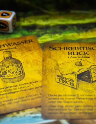 Zwei Spielkarten auf einem Tisch zeigen Illustrationen und Texte. Die linke Karte mit der Überschrift "Kirschwasser" zeigt eine gezeichnete Flasche Kirschwasser. Die rechte Karte mit der Überschrift "Schreibtisch-Blick" zeigt einen Schreibtisch mit einem Kuchen darauf. Im Hintergrund sind drei bunte Würfel sichtbar.