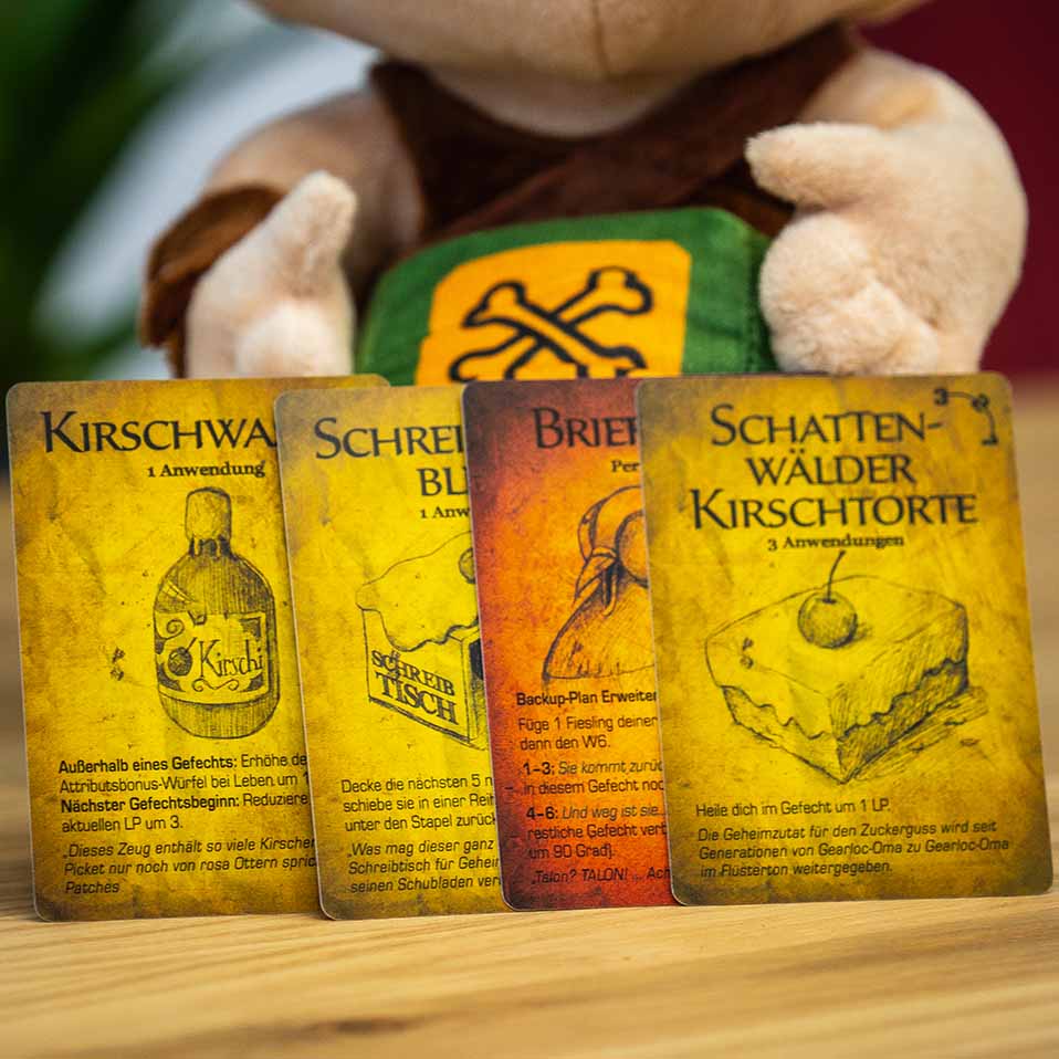 Vier bunte Spielkarten stehen nebeneinander auf einem Holztisch. Sie zeigen verschiedene Illustrationen: eine Flasche mit der Aufschrift "Kirschwasser", ein Tisch, ein großes Brötchen und ein Stück Kirschkuchen. Hinter den Karten ist ein Plüschtier mit brauner Kleidung und einem aufgestickten, gelben Symbol mit zwei gekreuzten Knochen zu sehen.