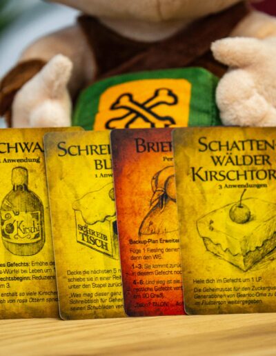 Vier Spielkarten mit verschiedenen Motiven und Texten liegen auf einem Holztisch. Hinter den Karten ist ein Stofftier mit einem grünen und gelben Muster auf der Brust zu sehen. Die Karten tragen die Titel "Kirschwasser", "Schreiberblut", "Brief" und "Schattenwälder Kirschtorte" und haben jeweils spezielle Anwendungsbeschreibungen und Illustrationen, die eine Flasche, eine Rolle Papier und ein Stück Torte zeigen. Die Details der Karten sind klar erkennbar, da sie im Vordergrund des Bildes stehen.