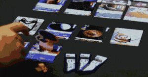 Eine Hand greift nach einer Karte auf einem Tisch mit mehreren ausgebreiteten Spielkarten. Die Karten zeigen Bilder von Planeten und anderen Weltraummotiven. Neben den Karten liegt eine kleine Spielfigur aus Holz. Es handelt sich um ein Strategiespiel mit einem Weltraumthema.