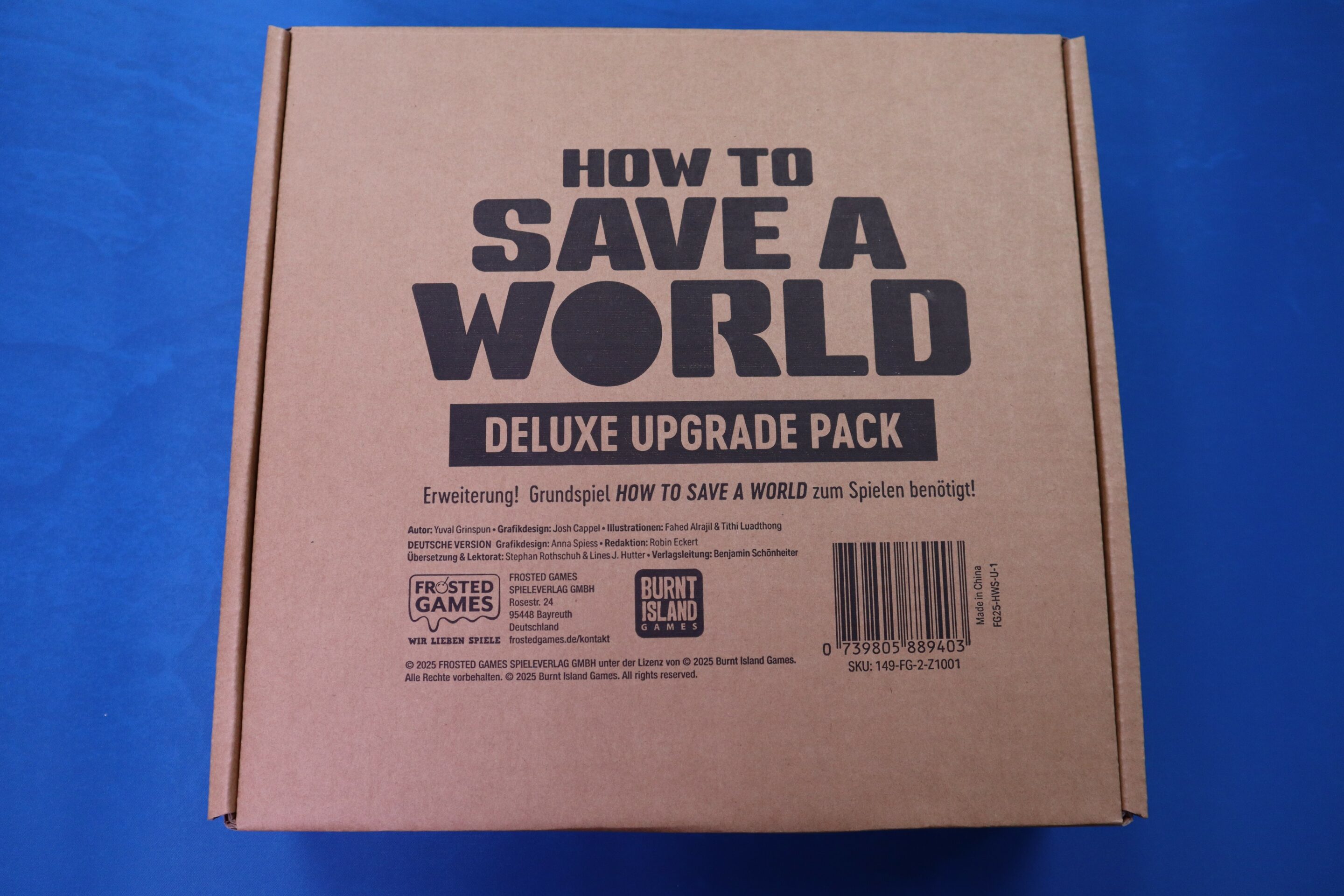 HTSAW Upgrade 1 Ein brauner Karton mit dem Text "How To Save A World: Deluxe Upgrade Pack" in schwarzer Schrift. Der Karton ist eine Erweiterung für das Grundspiel und enthält Details zu Autoren, Grafikdesignern und Illustratoren. Am unteren Rand befinden sich Logos von Frosted Games und Burnt Island Games sowie ein Barcode. Der Hintergrund ist blau.
