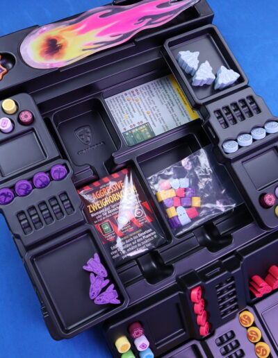 Nahaufnahme eines Brettspiel-Inlays aus schwarzem Kunststoff auf blauem Hintergrund. Das Inlay enthält verschiedene bunte Spielsteine und Marker in Fächern, darunter runde Knöpfe in mehreren Farben, violette Figuren und weiße Bausteine in Form von Tannenbäumen. In der Mitte ist ein Kartenspiel mit der Aufschrift "Aggressive Zweihörnchen" zu sehen, darunter ein transparentes Tütchen mit bunten, kleinen Spielensteinen. Oben links befindet sich ein Abzeichen mit einem flammenähnlichen Muster.