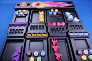 Ein Tischspiel-Organizer mit mehreren Fächern, gefüllt mit bunten Spielmarken und Figuren. Die Marken sind in verschiedenen Farben wie Gelb, Rot, Lila und Blau. Einige Marken haben Symbole und Muster, andere sind in Form kleiner Raumschiffe gestaltet. Der Organizer ist schwarz und hat Einteilungen für die verschiedenen Spielkomponenten. Im Hintergrund ist ein blaues Tischtuch sichtbar.