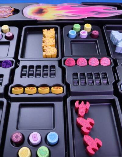 Ein Tischspiel-Organizer mit mehreren Fächern, gefüllt mit bunten Spielmarken und Figuren. Die Marken sind in verschiedenen Farben wie Gelb, Rot, Lila und Blau. Einige Marken haben Symbole und Muster, andere sind in Form kleiner Raumschiffe gestaltet. Der Organizer ist schwarz und hat Einteilungen für die verschiedenen Spielkomponenten. Im Hintergrund ist ein blaues Tischtuch sichtbar.