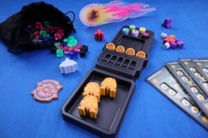 Brettspiel-Zubehör auf einem blauen Untergrund: Links ist ein schwarzer Stoffbeutel, aus dem bunte, durchsichtige Würfel in Rot, Grün, Blau und Schwarz herausgefallen sind. Rechts davon befindet sich eine schwarze Plastikbox mit gelben und orangen Spielmarken, darüber bunte runde Spielteile. Im Hintergrund liegen bunte hölzerne Spielfiguren und Karten mit Spielinformationen. Oben ist eine flammende Meteorfigur zu sehen. Links unten liegt ein Emblem in Form eines Abzeichens.