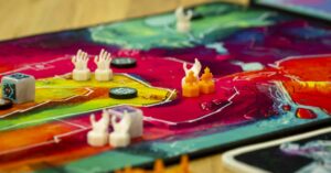 Nahaufnahme eines bunten Brettspiels mit abstrakten, farbigen Zonen. Auf dem Spielbrett sind Würfel, weiße Spielfiguren in Form von kleinen Händen und orangefarbene Figuren in Flammenform verteilt. Einige runde, schwarze Marker sind ebenfalls sichtbar. Die lebhaften Farben und unterschiedlichen Spielfiguren stehen im Fokus des Spiels.