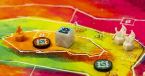 Nahaufnahme eines bunt gestalteten Spielbretts mit geometrischen Mustern. Darauf befinden sich mehrere Spielfiguren: ein oranger Flammenmarker, zwei weiße Handfiguren und ein weißer Würfel mit einem Symbol auf der oberen Seite. Zusätzlich sind zwei runde Markierungen mit einem Sternmotiv zu sehen. Die Farbgebung des Brettes reicht von Grün über Gelb bis Rot und enthält nummerierte Bereiche.