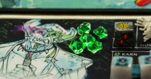 Ein Detailausschnitt eines Brettspiels zeigt eine illustriert dargestellte, skelettartige Figur mit Hörnern und ein Bündel von fünf grünen, kristallförmigen Spielsteinen. Daneben befindet sich ein mehrseitiger Würfel mit einem gelben Stern und zwei roten Totenköpfen als Symbole.