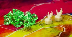 Haufen von grünen, kristallähnlichen Spielsteinen neben zwei weißen Spielfiguren in Form von Händen auf einem bunt illustrierten Spielbrett.