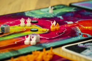 Ein farbenfrohes Brettspiel, auf dem mehrere detailreiche Spielfiguren und Spielsteine zu sehen sind. Im Vordergrund befinden sich verschiedene cremefarbene und orangefarbene Figuren, darunter welche in Form von aufrecht stehenden Händen. Auf dem Spielfeld sind zudem runde Spielsteine und ein Würfel platziert. Die Farben des Spielfelds changieren zwischen grün, pink, rot und blau, was dem Spiel eine lebendige Atmosphäre verleiht.